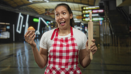 Woman holding muffin and rolling pin, wearing red checkered apron and grimacing in airport terminal...