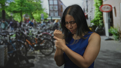 Fototapeta premium Woman clutching wrist on amsterdam street near parked bicycles, wearing blue dress and glasses; pain concern.