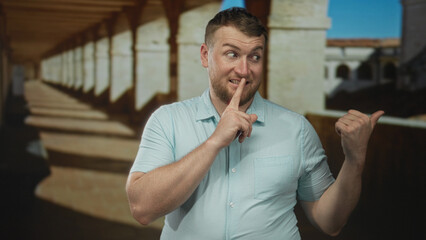 Naklejka premium Man with finger to lips shushing on an old town street beside stone colonnade, pointing thumb aside; secret mischief.