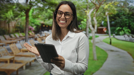 Woman holding tablet with both hands, smiling and tapping screen while standing beside wooden lounge chairs on a paved path near a building  joyful connection. © Krakenimages.com