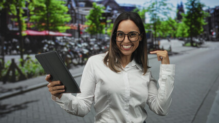 Fototapeta premium Young woman wearing white shirt and glasses holding tablet, hand touching screen and smiling on street lined with bicycles and trees; confidence joy.