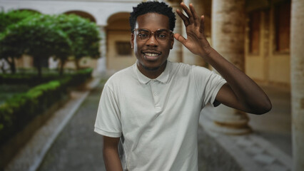 Fototapeta premium Man in white polo shirt with glasses gestures with raised hand in building; confidence focus optimism empowerment.