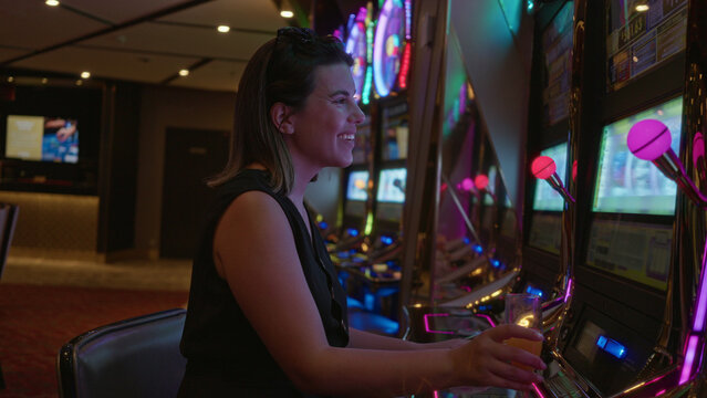 Woman holding a champagne glass drinking while playing slot machine in a casino building with neon lights; win joy.