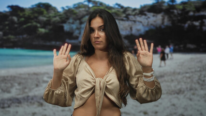 Woman hands raised palms out on beach showing bare navel and casual blouse, neutral expression and shrugged shoulders; indifference.