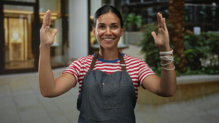 Naklejka premium Woman hands raised facing camera on a street in front of shops wearing apron and striped shirt; welcoming service.