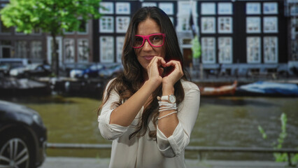 Fototapeta premium Woman wearing pink glasses forming a heart with hands on a street by an amsterdam canal, smiling; love connection.