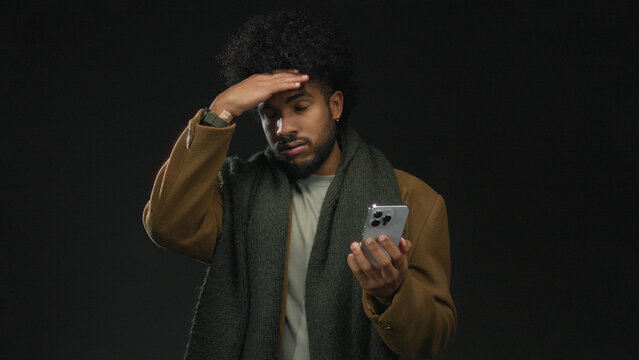 Man in winter attire looking at smartphone with a concerned expression against a black background, highlighting a moment of anxiety or surprise.