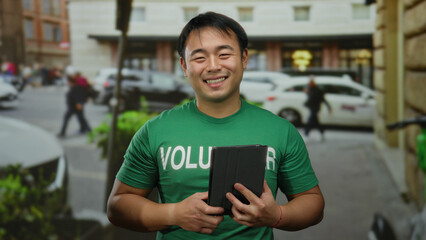 Fototapeta premium Young man holding tablet smiling in urban street with green shirt volunteering
