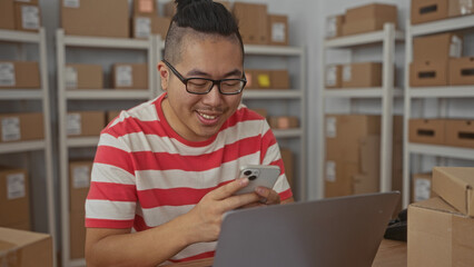 Man wearing glasses holding smartphone and typing with both hands beside laptop amid stacked parcel boxes on shelves in an office building, smiling; small business productivity.