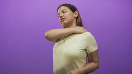 Obraz premium Woman rubbing her shoulder with hand, eyes closed, wearing yellow t shirt in purple studio; muscle tension discomfort.