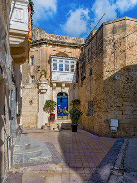 Attard, Malta &ndash; old town alley with light paving, white wooden balcony, blue door, potted plants and sandstone facades under blue sky