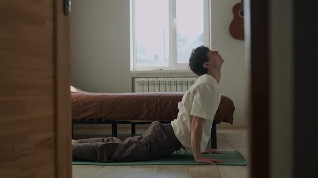 Beginner man doing simple yoga and morning stretching on mat in bedroom, casual home workout in daylight, behind open door view, real lifestyle routine