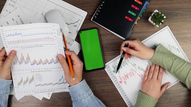 Professional team reviewing financial reports, working collaboratively with green-screen smartphone, notebook, spreadsheets on wooden workspace

