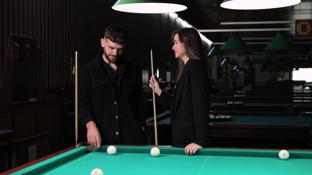 Attractive couple learning billiards together, boyfriend guiding girlfriend through game techniques and rules at stylish billiard club with warm lighting and green table
