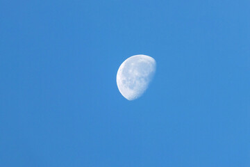 
This image captures a striking view of a waning gibbous moon suspended in a vast, clear blue sky during daylight hours. The lunar surface is rendered with impressive detail