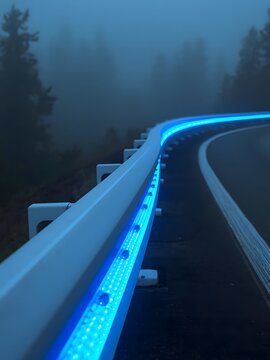 Curving highway guardrail illuminated by vibrant blue LED lights at dusk, shrouded in atmospheric fog, creating surreal and safe passage through misty forest road, adventure, journey, guidance.