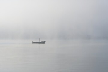Small wooden boat gliding through calm water in a foggy landscape, with mist enveloping the surroundings and creating a serene atmosphere