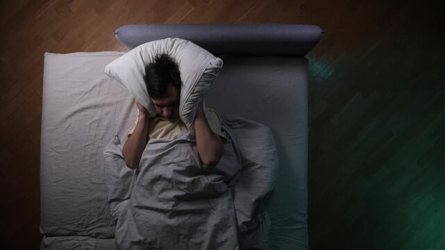 Man lying in bed overhead view, covering ears with a pillow, experiencing sleeplessness and frustration during the night due to noise disturbing his rest at bedtime