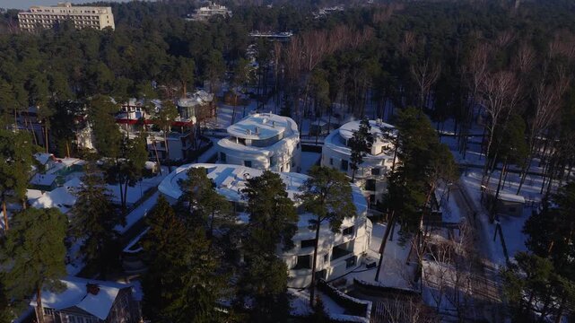 Aerial drone pans and descends over Jurmala, Latvia, showing rounded white villas, terraced circular roofs, winding roads, tall pines, and long winter shadows.