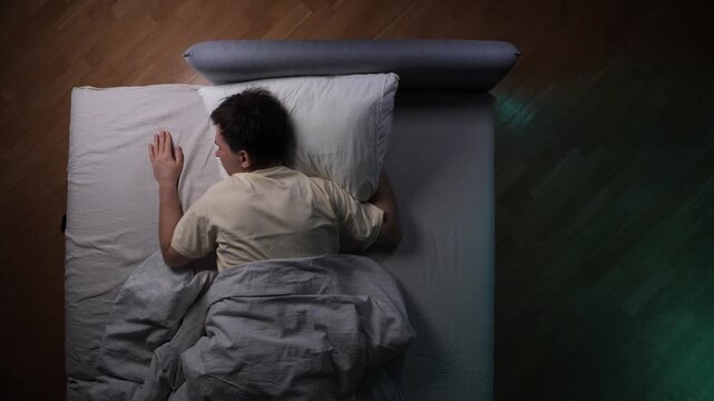 Reluctant man lying in bed, extending his arm to throw a ringing smartphone off the mattress, feeling annoyed and trying to avoid waking up in the morning