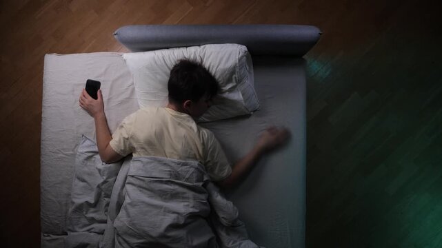 Man sleeping in bed with a phone in his hand, struggling with restlessness and insomnia due to screen time before bed, highlighting technology addiction and poor sleep habits