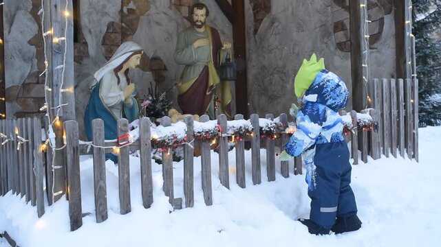 a little boy stands in front of a nativity scene in winter outside	