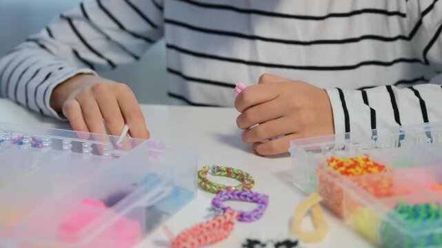 A girl creates a bracelet using colored rubber bands. She uses a weaving slingshot and a hook. Her workspace includes various rubber bands and tools. This is a crafting hobby