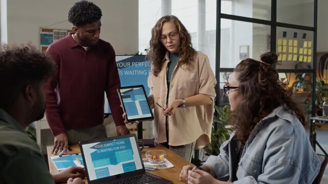 Handheld zoom out shot of group of four diverse designers using digital tablet and laptop to discuss ideas during meeting in bureau