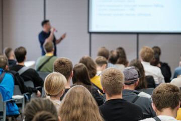 Fototapeta premium Live presentation in front of attentive audience in conference setting. Speaker explains slides and engages crowd through talk visuals in classroom style venue.