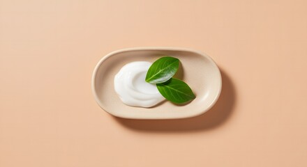 A small dish of white cream accompanied by two vibrant green leaves on a peach background