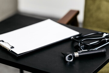 Medical examination tools including a stethoscope and otoscope on a black table with a blank clipboard ready for patient information in a clinical setting