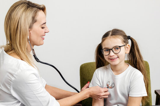 Female pediatrician using stethoscope to examine smiling girl with glasses sitting on a green chair in a bright medical office setting