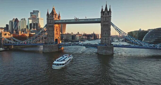 Evening Tower Bridge framed by London skyline at golden hour sunset, warm light reflecting on the Thames River, passenger boat cruises past historic towers and modern city skyscrapers. Aerial footage