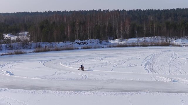 Aerial passes over a frozen lake in Latvia as a motorcyclist circles, carving spirals and skid marks. Birch and pine trees, reed beds, and a beached paddleboat are visible.