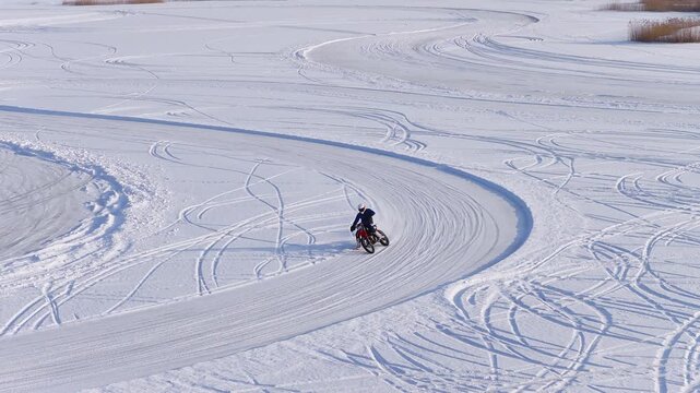Aerial view shows a motorcyclist drifting and turning on a frozen lake in Latvia, bright winter daylight, groomed icy circuit, reeds and buildings on the shore.