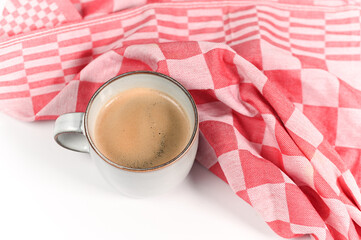 A cup of hot coffee on a white background with a red checkered kitchen towel. Cozy morning drink still life, minimal breakfast concept with copy space.