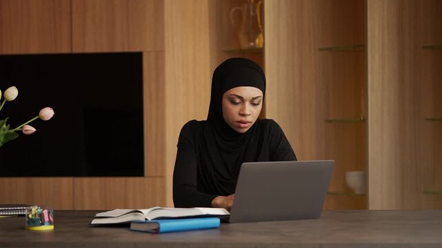 Focused Muslim woman in hijab studying online at home, e-learning concept