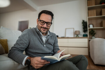 Fototapeta premium Smiling man reading book at home enjoying leisure time
