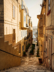 Obraz premium Sunlit narrow street with traditional stone architecture and harbor view in a Mediterranean town. birgu, Valetta. Malta