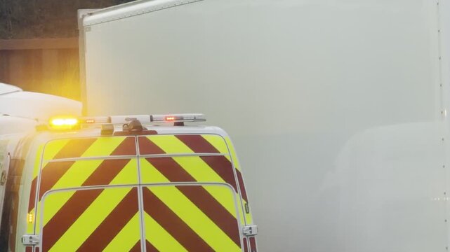 Highway maintenance van with flashing yellow and red beacons parked beside a white semi-trailer