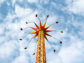 Vibrant image of a tall chair swing amusement ride, captured from below against a partly cloudy blue sky. Colorful chairs with silhouetted riders in motion, evoking feelings of summer, joy, and advent © Kristin