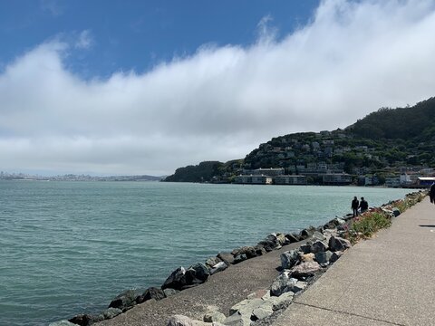 Coastal walkway lined with rocks and flowers along turquoise bay, hillside town of Sausalito in distance, and foggy clouds rolling over Marin headlands under blue sky