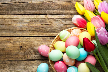 Painted Easter eggs in wicker basket and beautiful tulips on wooden table, flat lay. Space for text