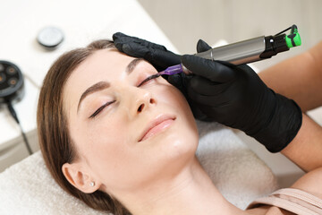 Naklejka premium Beautician making permanent eyeliner makeup to young woman in salon, closeup