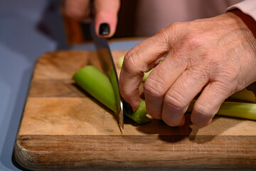 The hands of a mature woman with painted nails is cutting some celery on a well used wooden cutting board.  Close-up of a hand and knife.