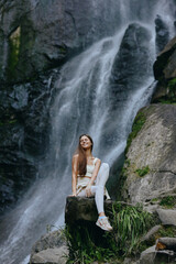 Obraz premium young woman sitting by waterfall in nature, wearing white clothes and sneakers, enjoying serene outdoor moment with greenery and rocks around, peaceful and relaxed atmosphere