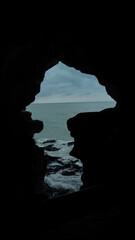 View of the sea through a dark coastal cave opening, dramatic rocky shoreline and waves under cloudy sky, mystery, freedom and nature exploration concept.