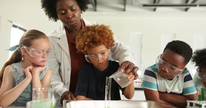 Teacher in lab coat placing cup, guiding teenager pouring into cylinder in lab forming green column