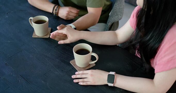 Couple holding hands at home, woman reaching across talking, mugs on coasters, smartwatch bracelets