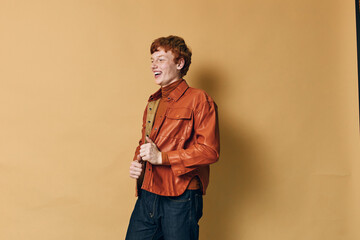 Young man with red hair wearing brown leather jacket and dark jeans, smiling and standing against...